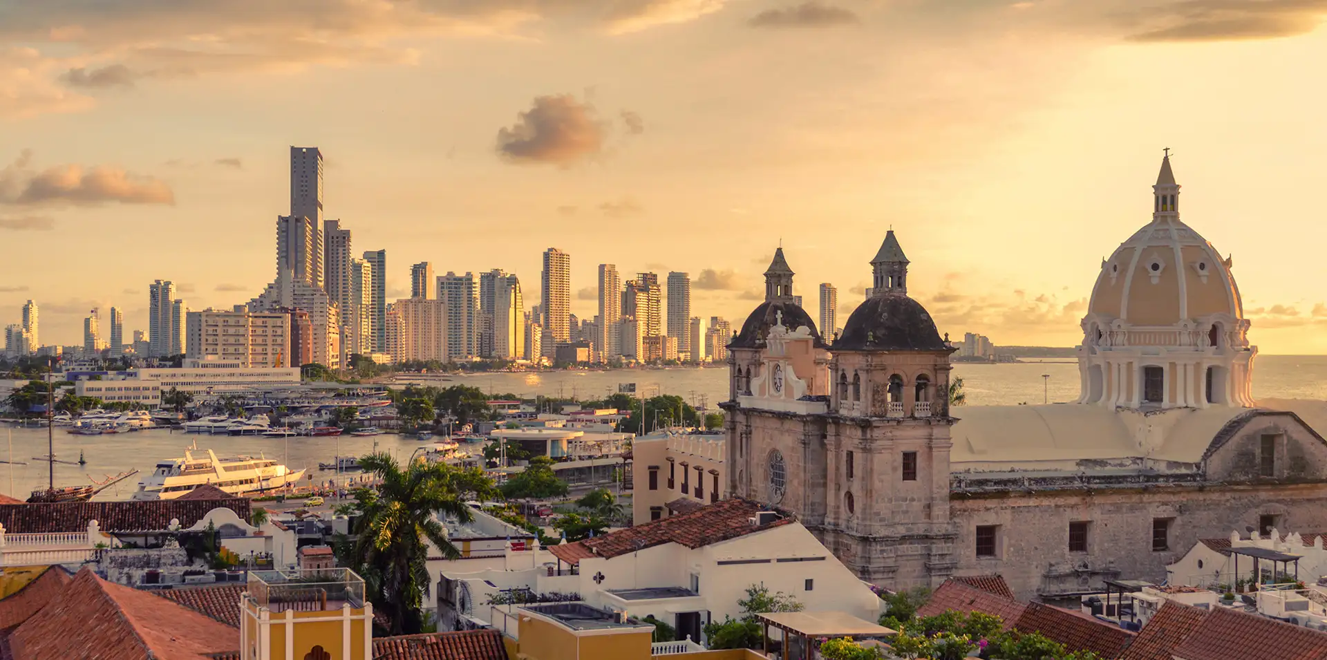 Panoramic sunset view of Cartagena’s historic center with cathedral domes in the foreground and the modern Bocagrande skyline by the Caribbean Sea.