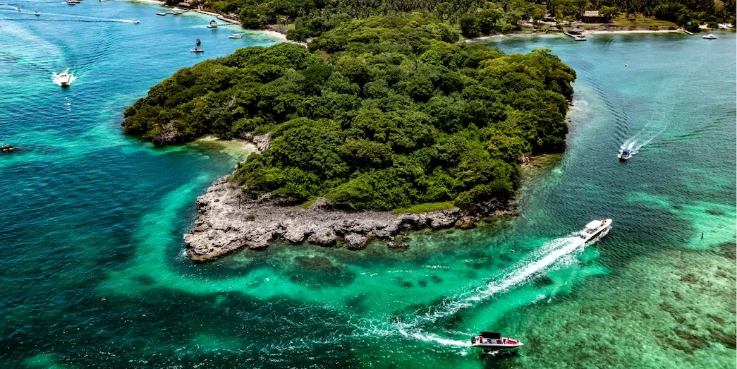 Aerial view of a small tropical island in the Rosario Islands with dense green vegetation, turquoise waters, and a boat leaving a white wake.