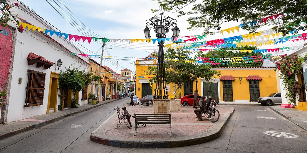 Colorful street scene in Getsemaní, Cartagena, with colonial houses, festive flags overhead, and a small plaza with benches.