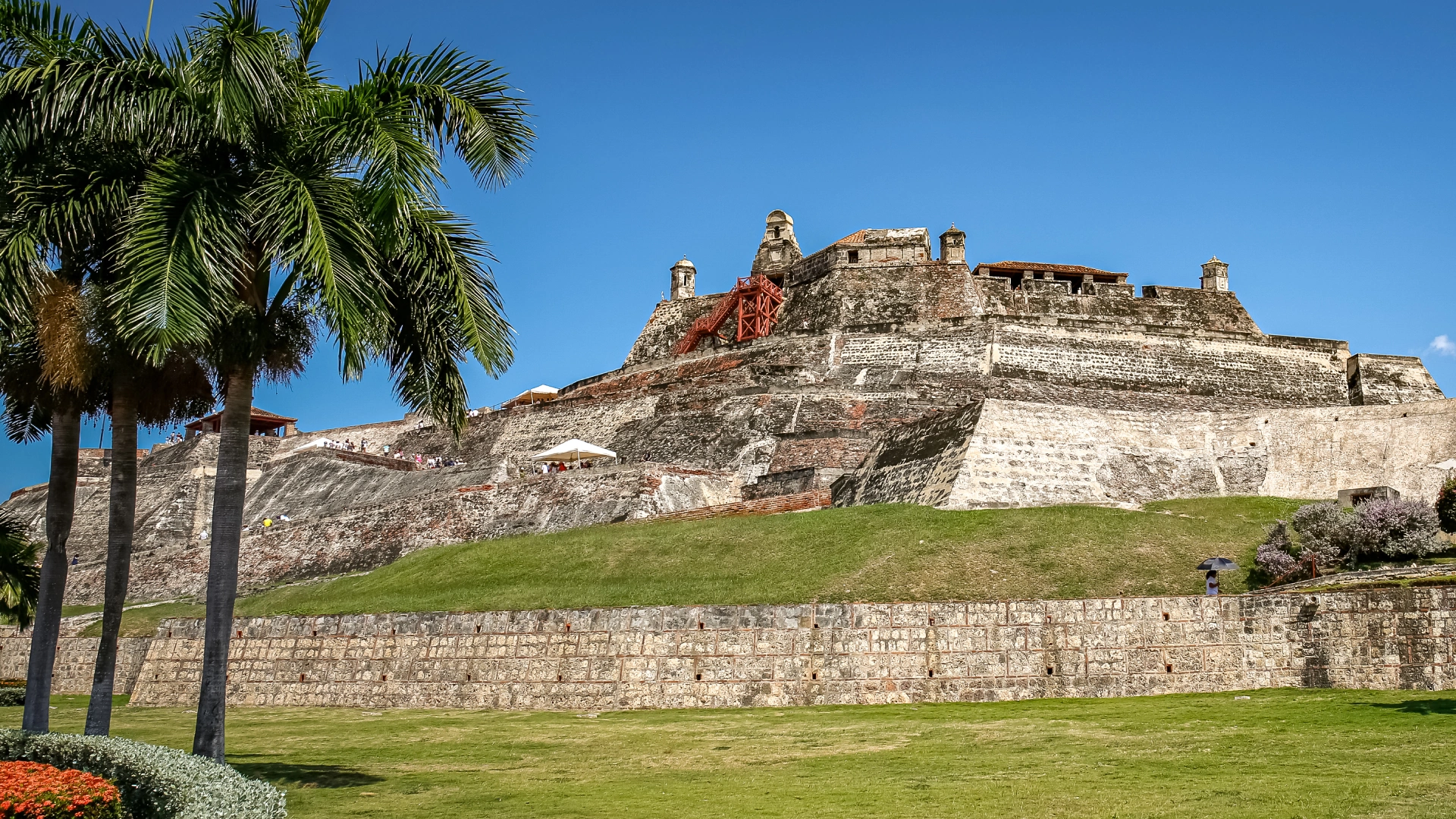 Castillo de San Felipe de Barajas rising above green lawns and palm trees under a clear blue sky in Cartagena, Colombia.